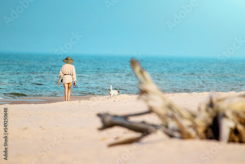 Plaża po której spaceruje Kobieta w kapeluszu, razem ze swoim psem. Widok na morze, ocean plaże i leżący konar drzewa.