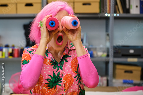 Seamstress with pink hair and colorfull clothes making funny faces with sewing thread reels in a sewing workshop.