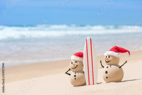 Two Sandy Christmas Snowmen are watching the waves, standing on beautiful beach with a surf board