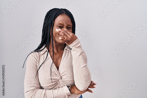 African woman with braids standing over white background smelling something stinky and disgusting, intolerable smell, holding breath with fingers on nose. bad smell