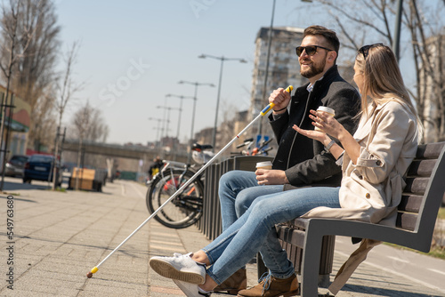 A blind male person using a white cane and sitting with his female friend on a bench. 