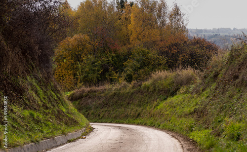 Kręta droga asfaltowa przebiegająca przez wzgórza pogórza Gór Świętokrzyskich .Szczyt jesieni . Drzewa w przepięknych barwach . Na tle szarego , zachmurzonego nieba . 