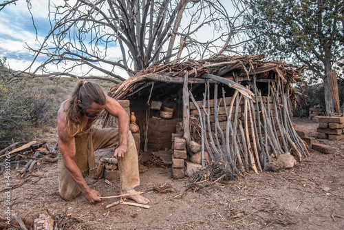 Man making friction fire in front of shelter