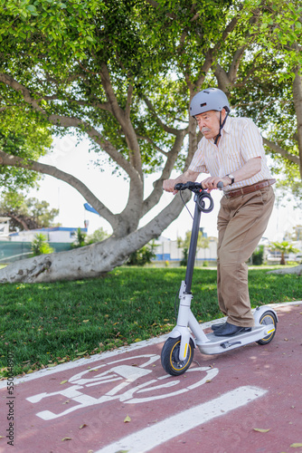 uomo anziano con il caschetto , monopattino elettrico corre con espressione impegnata in una pista ciclabile vicino a un parco cittadino .