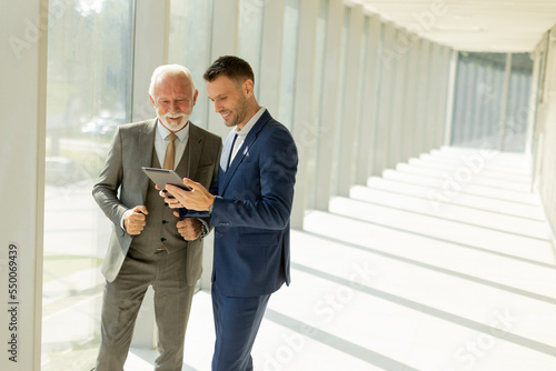 Mature businessman and his young colleague discussing finantial report on digital tablet in the office corridor