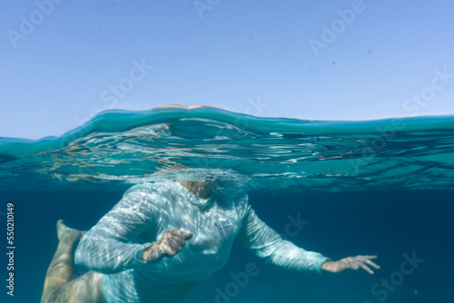 Underwater split shot of a woman wearing rashguard and swimming in the ocean, face is not visible only body. Water is deep green blue with blue sky showing on surface.