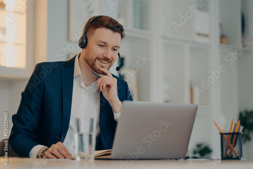 Professional male operator works in callcenter uses headset and laptop computer talks on video call or virtual webcam event wears formal clothes poses against cozy interior has online meeting