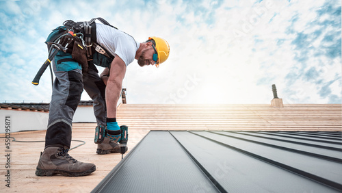 A roofer with a safety harness and tool belt is working with a electric screwdriver on the roof. He is anchoring the metal roofing with a screws.