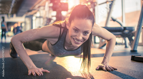 Fitness young woman doing push ups and looking at camera at gym. Friendly attractive girl doing push ups in front of sunlight at gym in the morning.