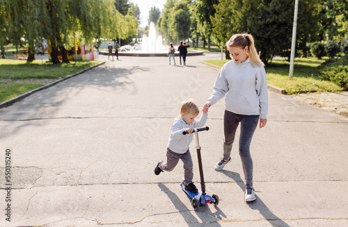 A boy with his mother riding in the park on a scooter