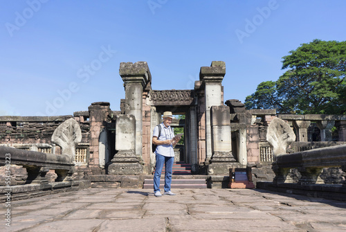 senior man with grey hair and beard,wearing hat,carry backpack and having camera, searching data on tablet at Phimai historical park,Thailand. concept ancient tourist, ecotourism, archeologist