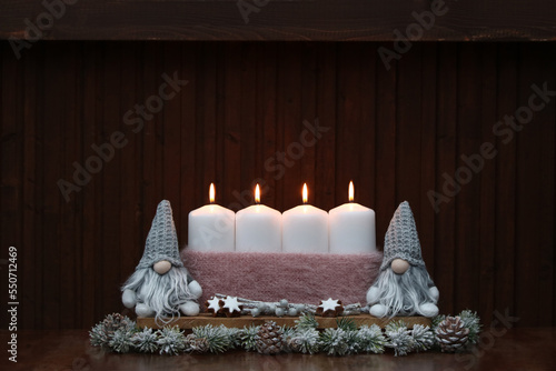Four white burning candles with Christmas santa clauses in front of a wooden wall with space for text.