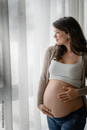 Portrait of a beautiful young pregnant woman standing by a big window and holding her belly. Always love your body.