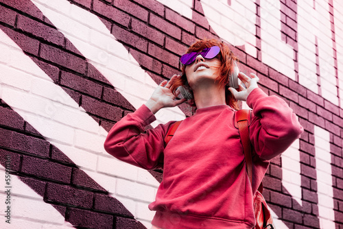 Stylish woman in magenta color jacket and heart shaped sunglasses wearing wireless headphones on her head and listening music. Urban city street fashion. Color of the 2023 year. Selective focus.