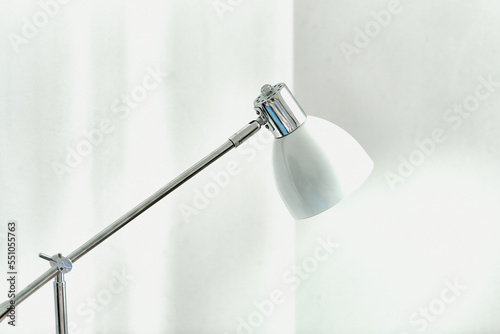 Chromowana lampa, lampka na stolik z białym kloszem na tle białych ścian rzucająca snop światła.
