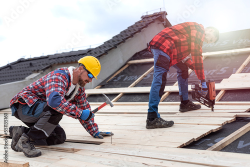 Two roofers at work. Nailing wooden battens on the roof with carpenter hammer or electric nailer.