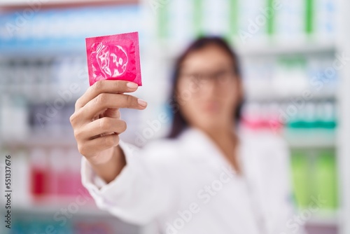 Young beautiful hispanic woman pharmacist smiling confident holding condom at pharmacy