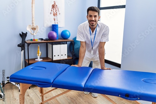Young hispanic man wearing physiotherapist uniform leaning on massage table at rehab clinic