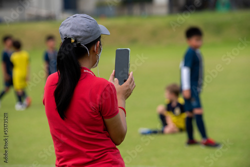 Mom standing and filming her son playing football in a school tournament on a sideline with a sunny day. Sport, outdoor active, lifestyle, happy family and soccer mom and soccer dad concept.