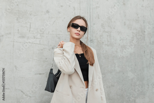 Stylish beautiful urban woman model with cool sunglasses in a fashionable beige blazer and a black top with a bag stands and poses near a gray concrete wall in the city