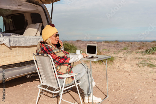 digital nomad woman works on laptop talking by mobile phone while traveling with camper van. Concept of modern people lifestyle