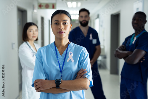 Portrait of biracial female healthcare worker with cancer ribbon in busy hospital corridor