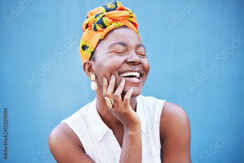 Happy black woman, african fashion and culture with turban scarf on blue wall background in Nigeria. Happiness, beauty and cultural style of young black person laughing with jewelry, makeup and smile