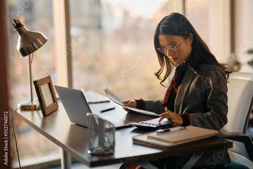 Asian female entrepreneur using calculator while going through business reports in office.