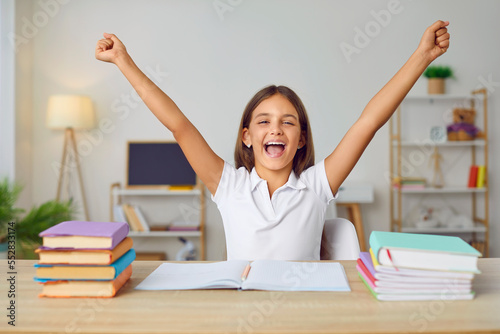 Happy, positive schoolgirl joyfully shouts, rejoicing that she has completed all her homework. Portrait of joyful girl sitting at home at table with textbooks and copybooks with raised hands.