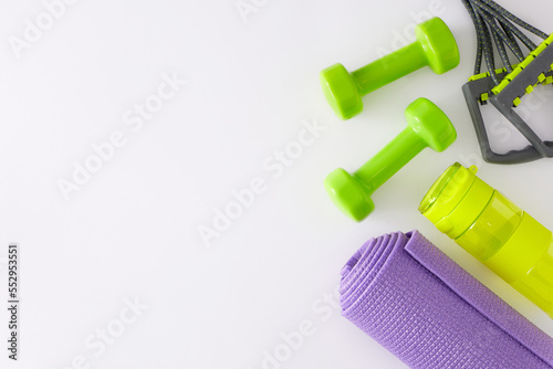 Fitness accessories concept. Flat lay composition of green dumbbells, violet exercise mat, bottle of water and elastic expander on white background with copy space. Minimal sport idea.