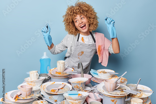 Positive woman dances carefree sings song foolishes around going to wash dirty dishes does chores cleans plates and cups does dishwashing at home wears striped apron and rubber gloves blue background