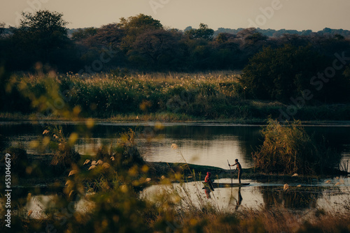 Idyllische Abendszene am Okavango - Zwei Jungen paddeln im Dämmerungslicht mit Einbaum Boot über den Fluss, Rundu, Namibia