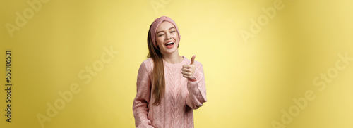 Happy amused beautiful young european girl in sweater, knitted headband showing good gesture, satisfied with service provided, winking sassy smiling with thumb-up in approval, giving recommendation