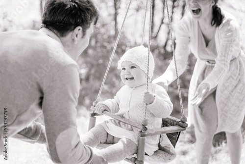 Stylish young parents swings their happy one-year-old daughter, on a wooden swing on a sunny day in the countryside, black and white image, the concept of child care, safety for children.