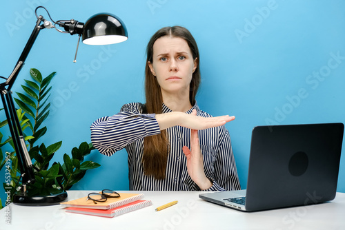 Strict employee young business woman 20s in shirt sitting work at white office desk with laptop holding stop gesture hands perpendicularly, posing isolated over blue color background wall in studio