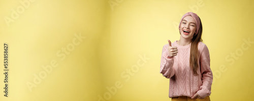 Happy amused beautiful young european girl in sweater, knitted headband showing good gesture, satisfied with service provided, winking sassy smiling with thumb-up in approval, giving recommendation