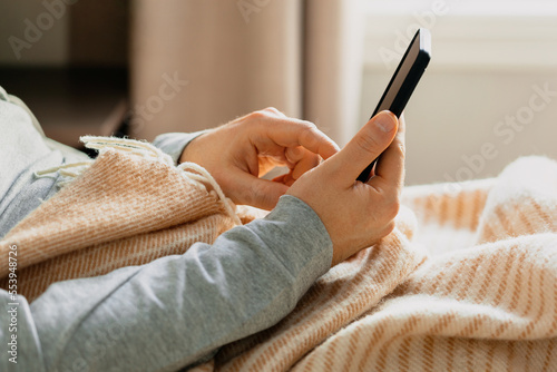 a caucasian man relaxing, using smart phone lying in bed under throw at home