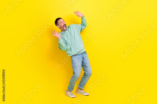 Full length photo of funky cheerful man look empty blank space ad poster raising up two arms isolated on yellow color background