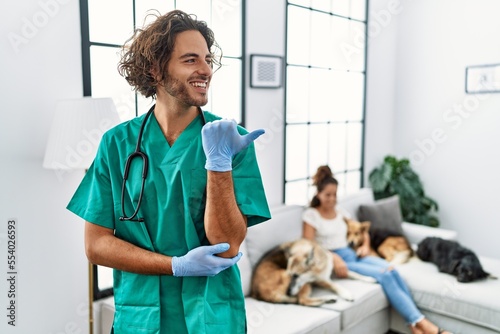 Young veterinarian man checking dogs at home smiling with happy face looking and pointing to the side with thumb up.