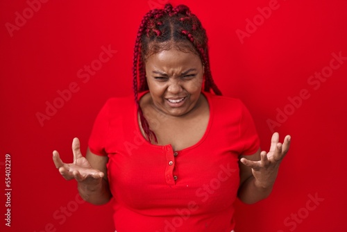 African american woman with braided hair standing over red background crazy and mad shouting and yelling with aggressive expression and arms raised. frustration concept.