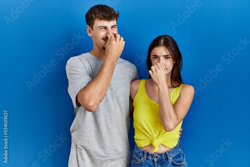Young hispanic couple standing together over blue background smelling something stinky and disgusting, intolerable smell, holding breath with fingers on nose. bad smell