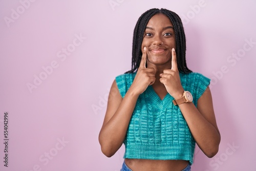 Young african american with braids standing over pink background smiling with open mouth, fingers pointing and forcing cheerful smile