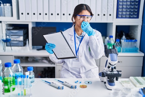 Hispanic young woman working at scientist laboratory smelling something stinky and disgusting, intolerable smell, holding breath with fingers on nose. bad smell