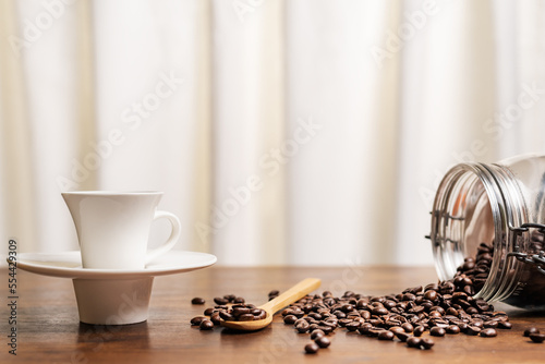 Cup of coffee next to an overturned jar with roasted coffee beans, some on a wooden spoon. Close-up.