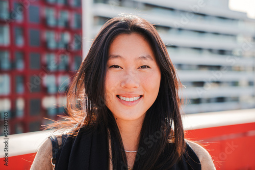 Close up portrait of asian young woman smiling and looking at camera with perfect white teeth, at the background city buildings. Attractive happy chinese female standing outdoors. High quality photo