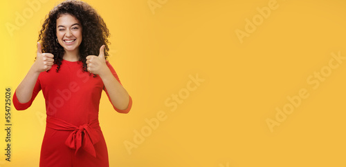 Creative and charismatic happy upbeat woman 25s with curly hair in red dress winking in approval and showing thumbs up with broad smile, satisfied giving positive reply over yellow wall