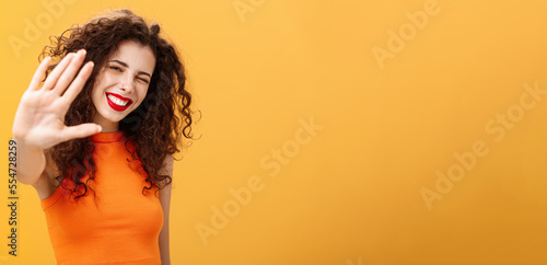 Give me five mate. Stylish friendly and joyful attractive woman with curly hairstyle winking and smiling happily pulling hand towards camera to greet or congratulate friend over orange background