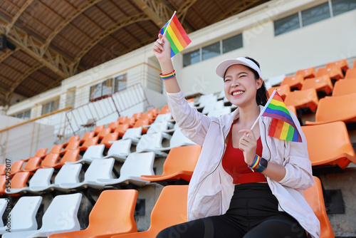 Happy and excited young female asian transgender LGBT sport fan cheering and watching the match with rainbow flag raising and wristband, she sitting amongst the rows of empty seats on stadium