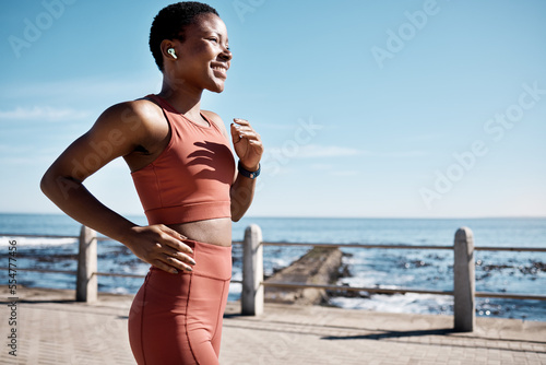 Black woman, music earphone or running by beach, ocean or sea in healthcare wellness or cardiovascular strength. Happy smile, sports runner or listening to workout training or marathon exercise radio