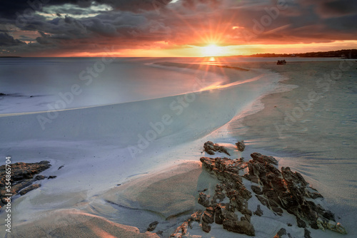 La Pelosa beach in Sardinia at sunrise with a sunburst and rocks in the foreground